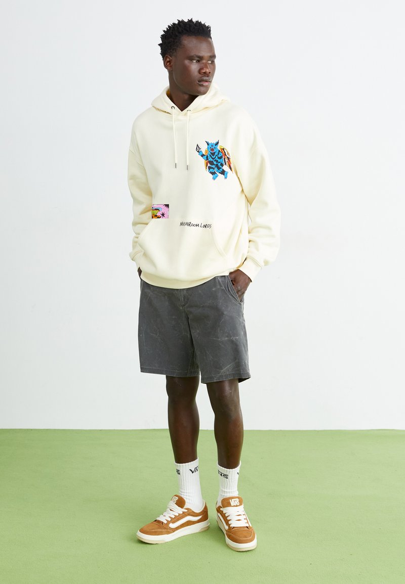 Kremowa bluza z kapturem z niebieską grafiką z przodu, szare szorty i beżowe sneakersy. Model stoi na zielonej podłodze na tle gładkiej białej ściany.