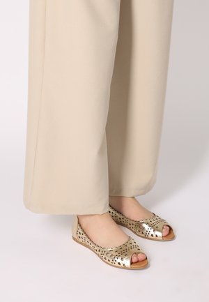 Gold perforated ballet flats with an open toe design, featuring triangle patterns and a flat sole, paired with beige wide-leg pants.