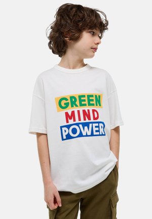 Garçon aux cheveux bouclés portant un t-shirt blanc avec le texte « GREEN MIND POWER » en lettres vertes, rouges et bleues, regardant sur le côté.
