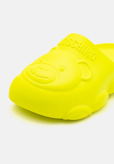 Zueco de goma amarillo con un diseño de cara de oso y logo grabado, que presenta una textura suave y forma redondeada. Diseño suave y ligero.