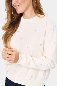 Šviesiai kreminė sportinė palaidinė su išsklaidytomis šviesiai mėlynomis širdelėmis, apvaliu iškirpimu, raukiniais ant rankovių ir prigludusia juosta. Minkštas audinio tekstūros.