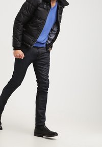 Schwarze, gesteppte Steppjacke, blauer V-Ausschnitt-Pullover, dunkle Jeans und schwarze Stiefeletten. Wirkt in Gehposition vor einem hellen Hintergrund.