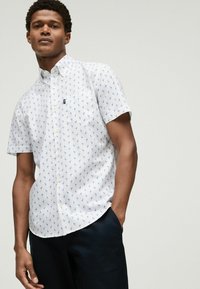 Camisa de botones de manga corta en blanco con estampado de flamencos azules, tejido de algodón texturizado, bolsillo en el pecho y dobladillo redondeado.