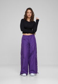 Schwarzes, langärmeliges Cropped-Top, kombiniert mit lockeren, lila Cargo-Hosen mit seitlichen Taschen, aus glattem Stoff, dazu weiße Sneakers.