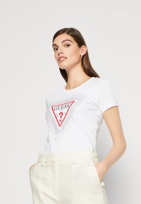 Hvid t-shirt med korte ærmer, der har et rødt trekantet logo med sølvnitter og ordet "GUESS" i fed, sort skrift. Glat stofstruktur.