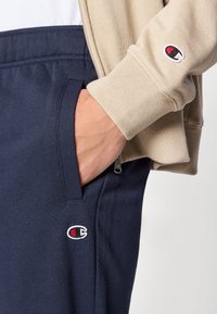 Pantalon de survêtement bleu marine avec poches latérales et logo Champion. Associé à une veste beige zippée. Tissu doux avec des poignets côtelés.