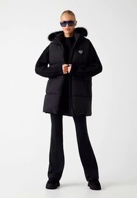 Gilet imbottito nero con maniche in maglia e cappuccio rifinito con pelliccia sintetica. Presenta una chiusura con zip e un dettaglio logo sul petto. Indossato sopra pantaloni neri.