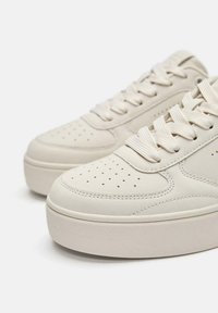 Adidași bej din piele cu vârf perforat, șireturi plate și talpă groasă de cauciuc texturată. Prezintă cusături contrastante de-a lungul marginii.