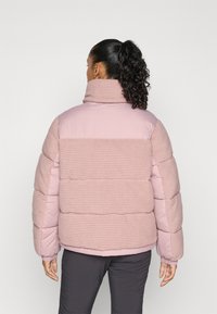 Person with curly hair in a high ponytail wearing a pink padded jacket with ribbed fabric panels, standing against a plain light background.
