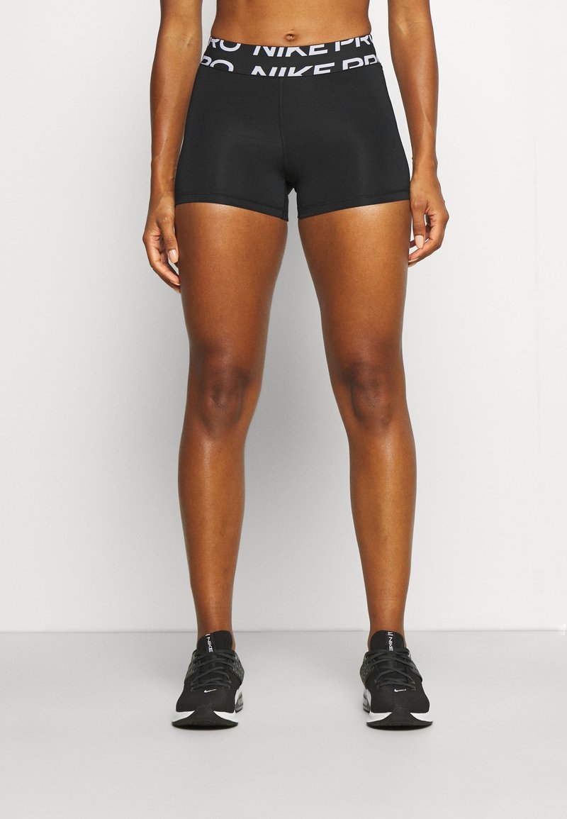 Personne athlétique portant un short Nike Pro noir et des chaussures de course Nike noires, debout devant un fond blanc uni.