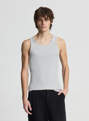 Młody mężczyzna z potarganymi brązowymi włosami, ubrany w szary bezrękawnik i czarne spodnie, stoi z jedną ręką w kieszeni.