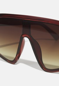Solbriller med et dristig, innhyllet design; rødtonede rammer med gradient brune linser. Har en glatt, blank overflate.