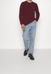 Bordeaux langemouw shirt met ronde hals, gecombineerd met blauwe rechte jeans en zwarte leren schoenen, wandelend op een lichte achtergrond.