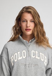 Grå sweatshirt med halvdragkedja. Har "POLO CLUB" i krämfärgad text och "Beyond Classics" broderat i grått. Täckande tyg.