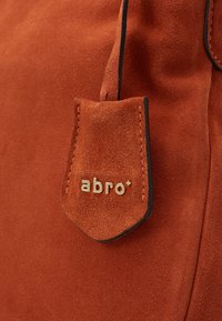 Oranssin mokkanahkalaukun yksityiskohta, jossa on läppä, jossa on kultainen "abro+"-logo ja kontrastiväriset mustat ompeleet reunoilla. Pehmeä tekstuuri on nähtävissä.