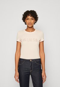 Camiseta ajustada de color rosa claro con mangas cortas, que presenta el logotipo "GUESS LOS ÁNGELES" en acentos brillantes. Combinada con jeans de mezclilla azul oscuro.