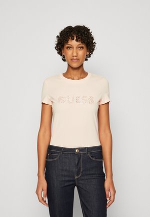 T-shirt ajusté rose clair à manches courtes, arborant le logo "GUESS LOS ANGELES" en accents brillants. Associé à un jean en denim bleu foncé.