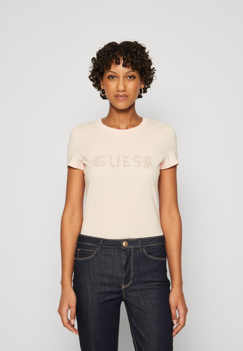 Camiseta ajustada de color rosa claro con mangas cortas, que presenta el logotipo "GUESS LOS ÁNGELES" en acentos brillantes. Combinada con jeans de mezclilla azul oscuro.