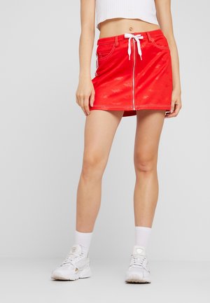 Femme portant une mini-jupe rouge zippée avec un cordon blanc, des chaussettes blanches à la cheville, des baskets blanches et un top court blanc côtelé.