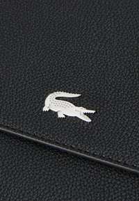Cuir texturé noir avec un détail logo crocodile argenté. Le design présente une finition lisse et brillante ainsi qu'une texture élaborée.