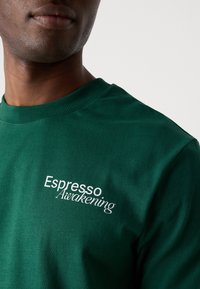 Green crewneck sweatshirt with ribbed collar. Features white embroidered text "Espresso Awakening" on the left side of the chest.