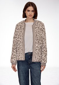 Jeune femme portant une veste zippée beige et marron à imprimé léopard sur un t-shirt blanc et un jean bleu, debout devant un fond blanc.