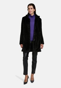 Black faux fur coat with a shawl collar, button closure, and two front pockets, worn over a purple ribbed turtleneck and black leather pants.