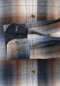 Kariertes Baumwollhemd in Blau-, Grau- und Beigetönen. Es hat einen knopfbaren Manschetten mit zwei braunen Knöpfen und eine Brusttasche.