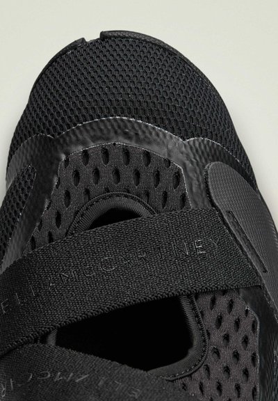 Black athletic shoe featuring mesh material with perforations, a smooth rubber toe cap, and Velcro strap detail. Textured accents throughout.