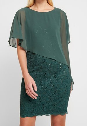 Kobieta w ciemnozielonej sukience z cekinową koronkową spódnicą i asymetryczną nakładką z muślinowym rękawem pelerynkowym.