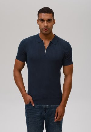 Polo shirt bleu marine à manches courtes en tissu côtelé avec un col zippé et une coupe ajustée, associé à un jean bleu, sur un fond neutre.