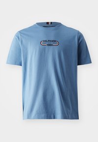 Jasnoniebieska koszulka z krótkim rękawem z napisem "Hilfiger New York City 1985" oraz czerwoną, białą i niebieską paskowaną strukturą na klatce piersiowej.