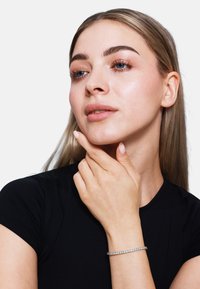 Sidabrinis apyrankė su apvaliais skaidriais akmenimis. Modelis su ilgais šviesiais plaukais vilki juodą prigludusį viršų, mąsliai laikydama smakrą. Taikytas neutralus makiažas.