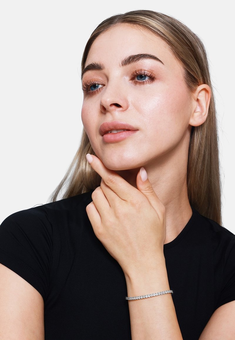 Sidabrinis apyrankė su apvaliais skaidriais akmenimis. Modelis su ilgais šviesiais plaukais vilki juodą prigludusį viršų, mąsliai laikydama smakrą. Taikytas neutralus makiažas.