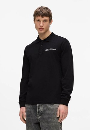 Mladý muž nosiaci čierne polo tričko s dlhým rukávom s logom "KARL LAGERFELD JEANS" a vyblednuté sivé džínsy, stojaci pred obyčajným pozadím.