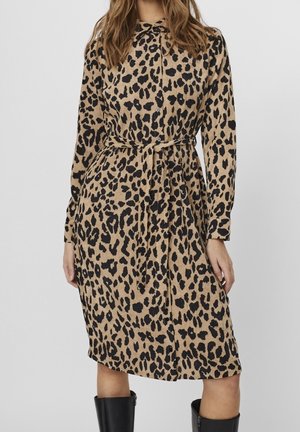 Kobieta ubrana w sukienkę z długim rękawem o długości do kolan w panterkę, z przodu na guziki i paskiem przewiązanym w talii, zestawiona z czarnymi, sięgającymi do połowy łydki butami.