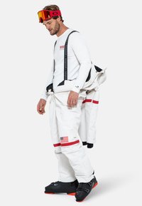 Traje de esquí blanco con detalles en rojo y negro, con cremalleras, correas y una parche de la bandera americana. Botas de esquí negras con detalles en naranja.