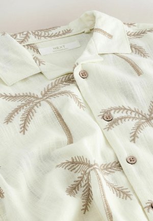 Chemise en lin blanche avec motifs brodés de palmiers beiges, boutons marron et col plié, étalée à plat.