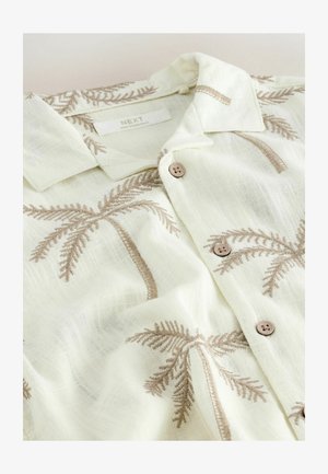 Weißes Leinenhemd mit beigen, bestickten Palmenmustern, braunen Knöpfen und einem umgeschlagenen Kragen, flach liegend.