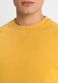 Sweat-shirt jaune à col rond avec un col côtelé, présentant une texture douce et un détail de couture subtil sur les coutures des épaules.