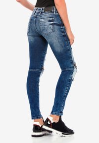 Blaue Skinny-Jeans aus Denim mit Reißverschlüssen an den Knien, einer leichten Verwaschung, fünf Taschen und einem strukturierten Bund. Getragen mit schwarzen Sneakers.