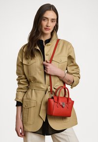 Piccola borsa a tracolla in pelle rossa con design strutturato, hardware in argento e manici superiori doppi. Il modello indossa un giaccone beige e una maglietta nera.