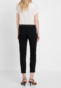 Kobieta stojąca tyłem, ubrana w czarne dopasowane spodnie z ćwiekowanym paskiem, białą bluzkę z krótkim rękawem i czarne sandały na obcasie.