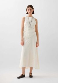 Cremfarbendes, ärmelloses Maxikleid mit strukturiertem Stoff und asymmetrischem Saum, kombiniert mit schwarzen flachen Schuhen und einer schlichten Halskette.
