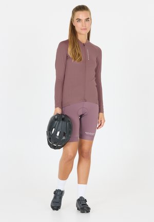 Das Radfahr-Outfit besteht aus einem helllila Langarmtrikot und passenden Shorts. Ein schwarzer Helm und Radsportschuhe mit einem Schnallenverschluss werden in der Hand gehalten.