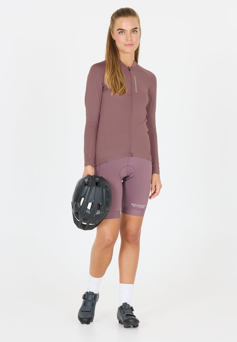 De fietsoutfit bestaat uit een lichtpaarse lange mouw trui en bijpassende shorts. Een zwarte helm en fietsschoenen met een gesp worden in de hand gehouden.