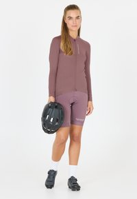 Vrouw staand in een mauve fietspak, met een zwarte fietshelm in haar hand, witte sokken en zwarte fietsschoenen, tegen een witte achtergrond.