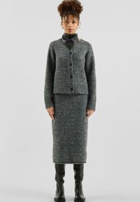 Frau in einer grauen gestrickten Strickjacke mit Knöpfen und passendem langen Rock, kombiniert mit schwarzen Lederstiefeln und einem Halstuch, vor einem schlichten Hintergrund stehend.