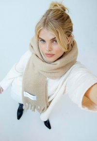 Écharpe beige en matériau doux, drapée autour du cou, avec des franges aux extrémités et une étiquette. Pull blanc texturé et bottes noires à bouts pointus.