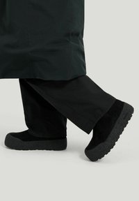 Mustat mokkanahkakengät, joissa on paksut harmaan tekstuuriset pohjat. Yhdistetty löysiin mustiin housuihin, joissa on vähäinen säde nilkan ylle.